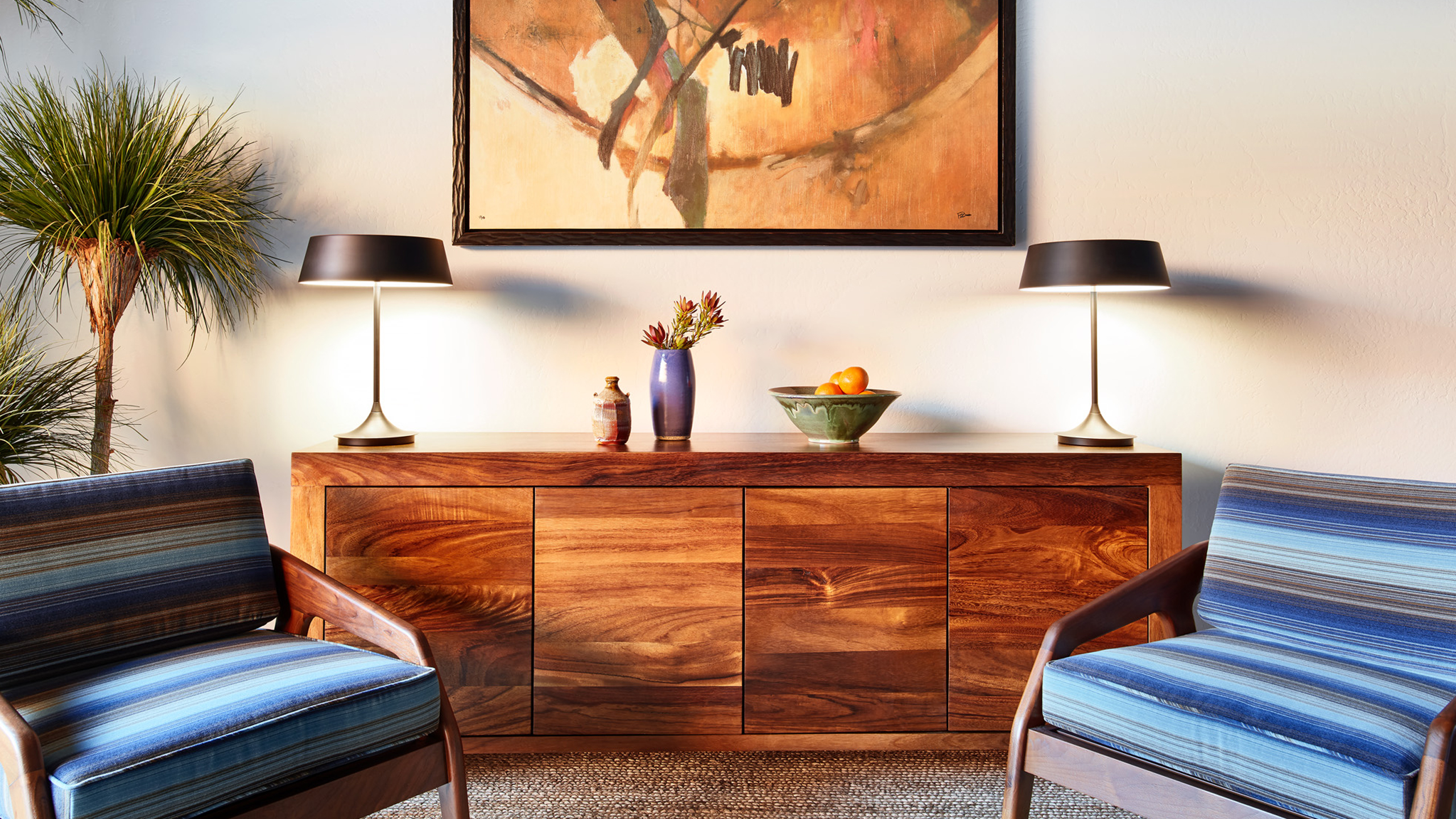 A lobby with a wooden sideboard, two lamps, a vase with flowers, a fruit bowl, and two chairs.