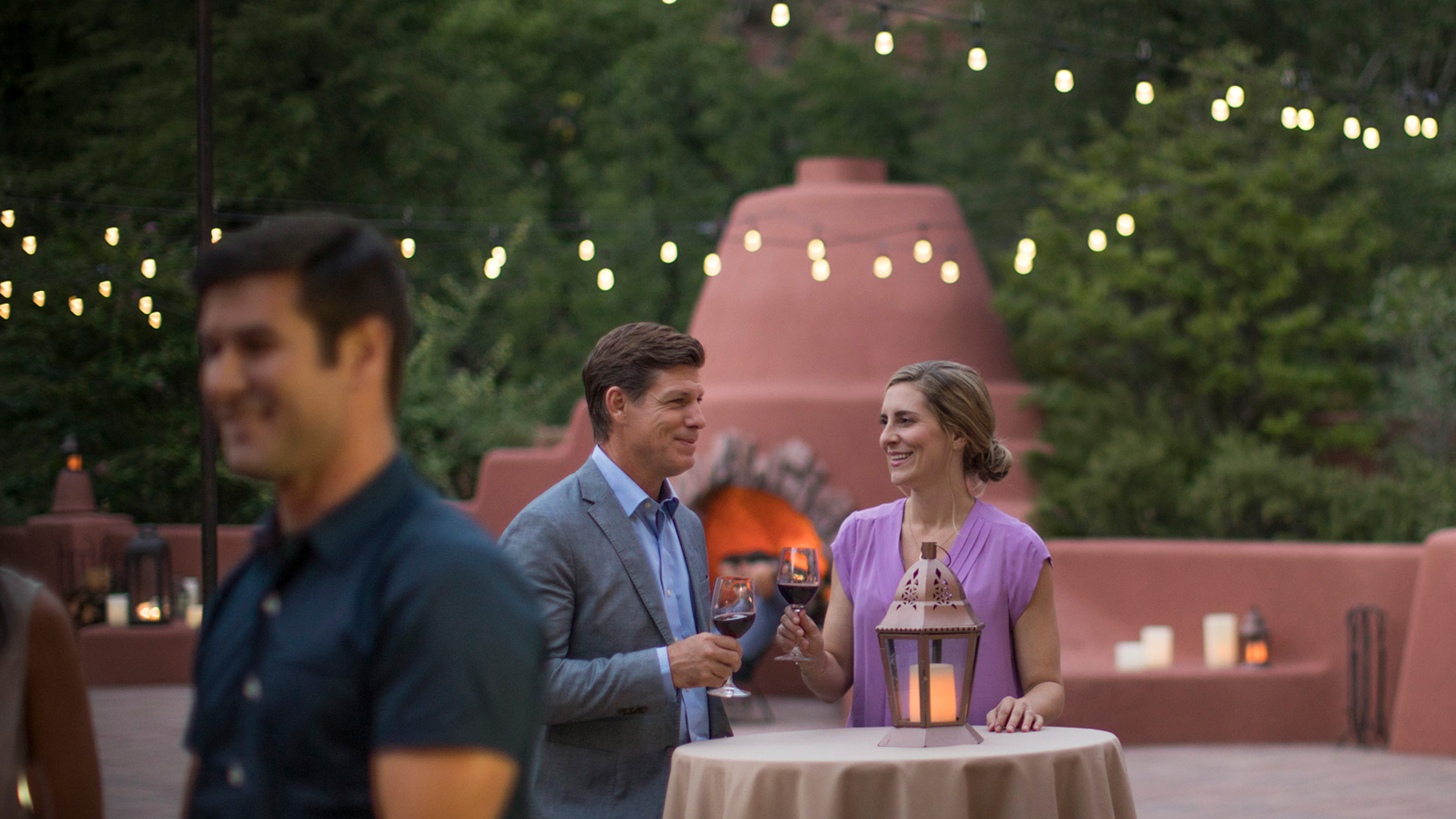 A man and a woman chatting at a standing table with a lantern on it near an adobe fireplace with string lights hanging above