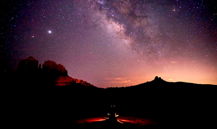 hiker sitting on trail at dark with sky filled with stars