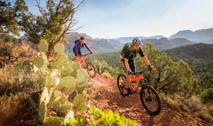 two mountain bikers - one female and one male - riding on trail in sedona with prickly pear cactus