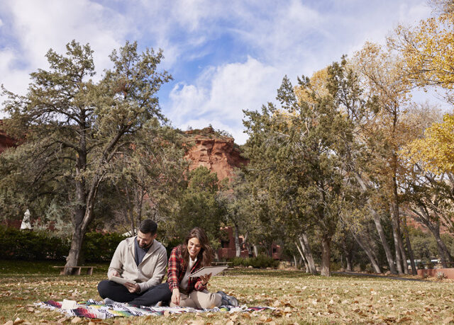 two people sitting in the grass on a blanket sketching in nature